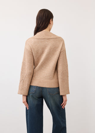 A woman viewed from the back wearing a beige knit cardigan with textured cable detailing on the sleeves and a wide ribbed collar, paired with dark blue jeans, against a plain light gray studio background.
