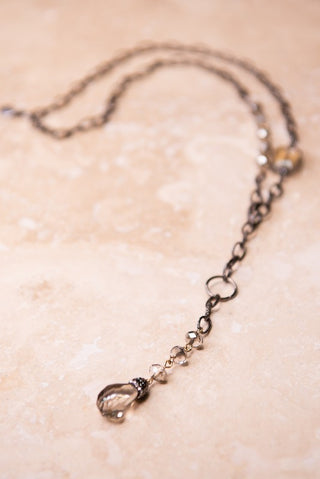 A delicate dark metal chain necklace with a small clear faceted bead and a tiny gold-toned charm, displayed on a textured beige stone surface.