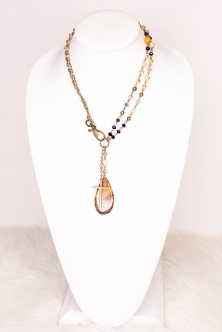 A multi-strand necklace displayed on a white mannequin bust. The necklace features a gold-tone chain, a strand of small black beads, and a central gold-tone pendant containing a pale pink stone and a small cross.