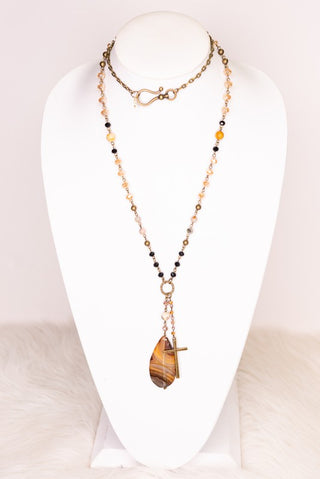 A long beaded necklace displayed on a white mannequin bust. The necklace features a layered design with a gold-tone chain, small black and cream beads, and a large tiger's eye pendant.