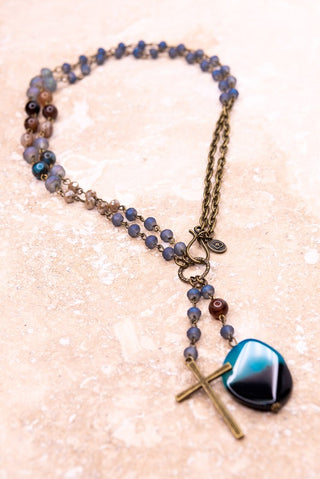 A beaded rosary-style necklace with blue and purple beads, an antique bronze cross, and a large iridescent blue and black heart-shaped bead, displayed on a light beige stone surface.