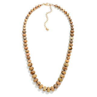 A beaded necklace made of flat, round natural stone beads in earthy tan, brown, and cream tones, with a gold-tone extender chain and clasp, shown on a plain white background.