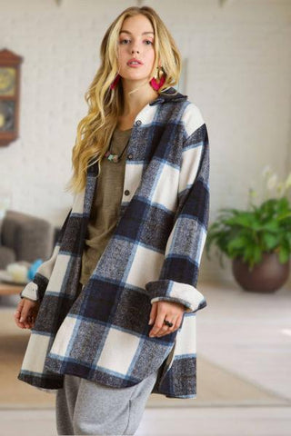 A woman wearing a large navy, white, and gray plaid hooded cardigan over a brown top and gray sweatpants, standing in a brightly lit indoor room with a plant in the background.