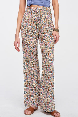 A model wearing high-waisted, wide-leg pants with a colorful geometric pattern on a beige base, paired with a blue top and brown sandals against a light gray background.