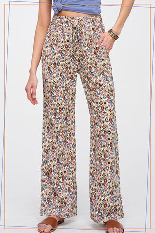 A person wearing loose, wide-leg pants with an all-over small geometric print in beige, brown, blue, and pink, paired with a blue top and brown sandals against a plain light background.