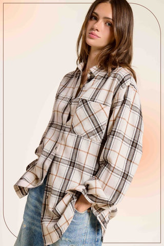 A woman wearing an oversized long-sleeve plaid button-up shirt in beige, brown, and black, paired with blue jeans, set against a soft beige background with a warm orange glow.