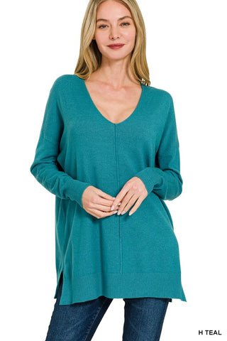 A woman modeling a teal long-sleeve knit tunic sweater with a V-neck and side slits, paired with blue jeans against a plain white background.