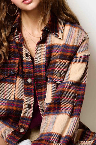 A close-up of a woman wearing a multicolor plaid button-up shirt jacket with beige, red, blue, and brown tones, layered over a dark purple top.