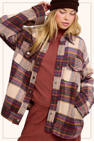 A woman wearing a plaid button-up flannel shirt jacket in beige, brown, and purple tones, layered over a rust-colored knit sweater and matching pants, with a dark brown beanie. The photo is cropped from the chest up against a plain light background.