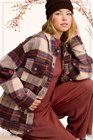 A woman models a long-sleeve button-up plaid overshirt in beige, brown, and burgundy tones, worn over a rust-colored corduroy jumpsuit with a dark brown beanie.