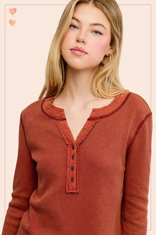 A woman wearing a rust-orange long-sleeve henley top with a waffle texture, photographed against a solid pale pink background.