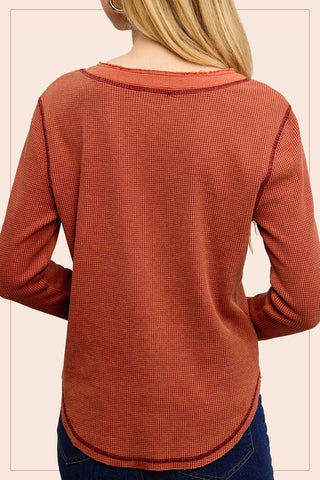 A woman shown from the back wearing a rust-orange ribbed long-sleeve top and dark blue jeans against a pale pink background.