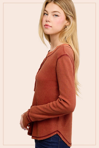 A blonde woman shown in side profile wearing a textured, rust-orange long-sleeve top and dark blue jeans against a solid pale beige background.