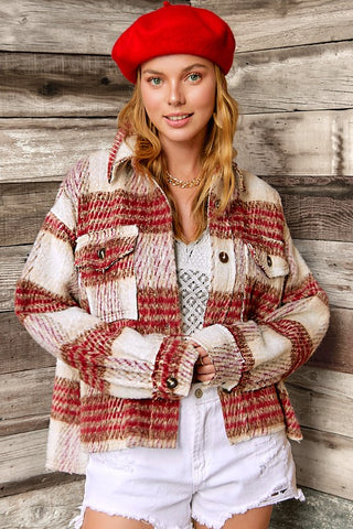 A woman wearing a chunky knit plaid jacket in red, beige, and white, paired with a red beret, white top, and white denim shorts, standing against a rustic wooden wall.
