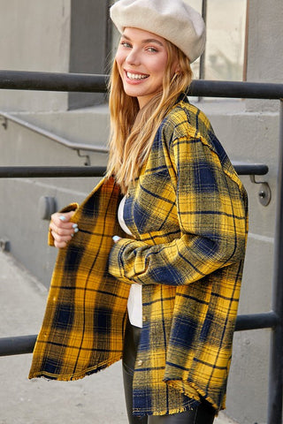 A woman wearing a yellow and navy blue plaid fringed cardigan, a cream beret, and a white top, standing outdoors in front of a gray concrete wall and black metal railing.