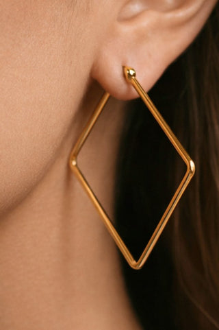 A close-up of a person's ear wearing a large open-frame geometric hoop earring in polished gold.