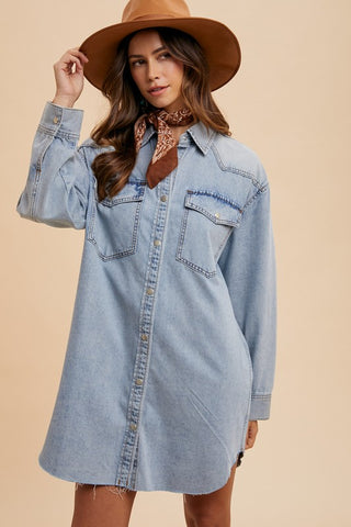 A woman wearing a light blue denim shirt dress with a brown wide-brim hat, a brown bandana, and brown boots, posed against a solid beige background.