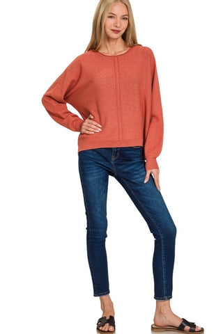 A woman modeling a loose-fitting, long-sleeve crewneck sweater in a heathered terracotta or burnt orange color, paired with dark blue skinny jeans and black slide sandals, against a plain white background.