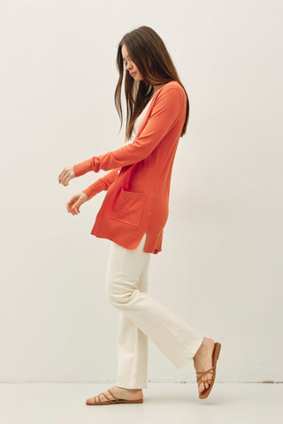 Woman wearing an orange long-sleeve top and white pants on a plain background