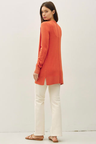 Woman wearing an orange long-sleeve top and white pants on a plain background