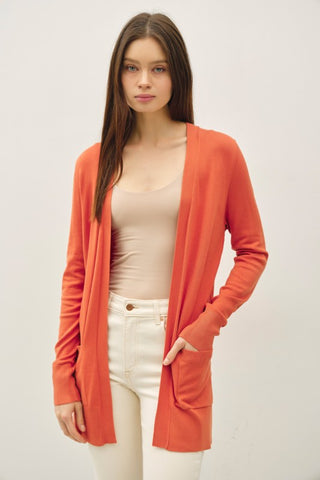 Woman wearing an orange cardigan over a beige top and white pants on a plain background