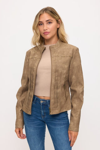 A woman modeling a cropped, long-sleeve brown faux-leather jacket with vertical pleat details on the front, worn over a beige ribbed crop top and blue jeans against a plain white background.