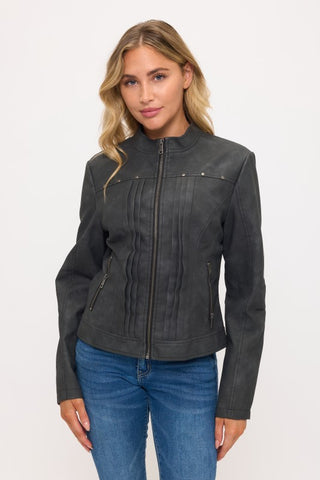 A woman wearing a dark gray faux-leather moto jacket with vertical pleats and zippered pockets, paired with blue jeans, against a plain white studio background.