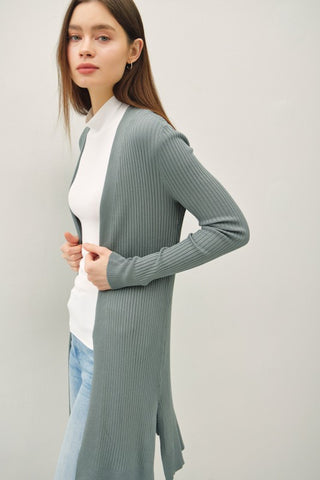 Woman wearing a long green cardigan over a white shirt and blue jeans on a plain background