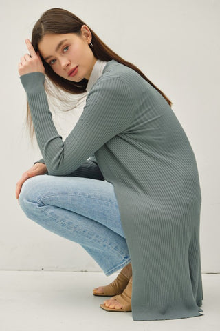 Woman wearing a long ribbed sweater and jeans sitting on the floor against a white background