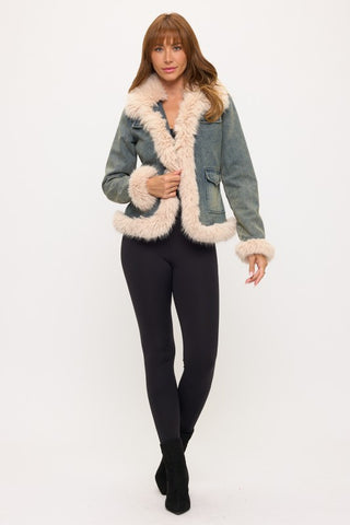 A woman modeling a light blue denim jacket with thick cream shearling-style trim and lining, paired with black leggings and black ankle boots, against a plain white studio background.