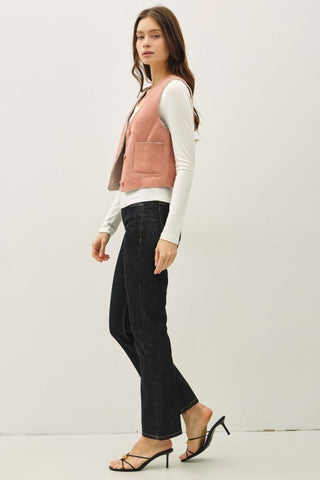 Woman wearing a pink vest over a white shirt with black pants on a plain background