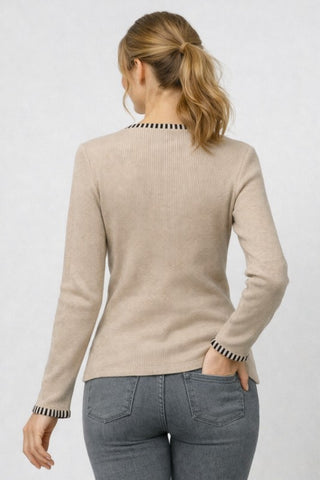 A woman viewed from the back wearing a light beige ribbed knit sweater with black and white striped trim at the crew neck and cuffs, paired with gray jeans, against a light gray studio background.
