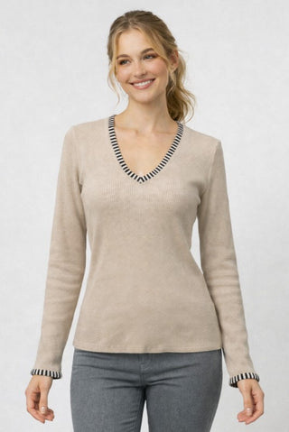 A woman wearing a light beige ribbed knit sweater with black and white striped trim along the V-neck and cuffs, paired with gray pants, against a light gray studio background.