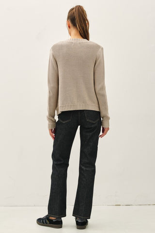 Person wearing a beige sweater and black pants against a white background