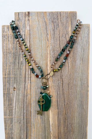 A beaded necklace with a green stone pendant and key charm, displayed on a rustic wooden stand against a white background.