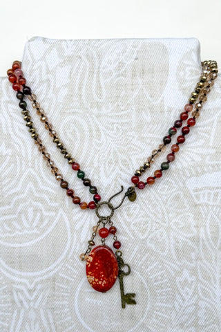 A bohemian-style multi-strand beaded necklace displayed on a white patterned fabric stand. The necklace features strands of red, brown, and dark green beads with a central antique bronze clasp holding a large oval red glass pendant decorated with gold glitter, and a small vintage-style metal key charm.