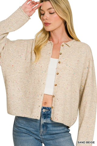 A woman wearing a cropped, long-sleeve knit cardigan in a heathered sand beige color with tiny multicolor speckles, layered over a white tank top and blue jeans. The image is cropped from the chest up against a plain white background.