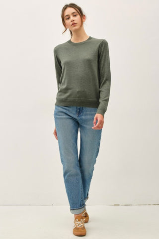 A woman modeling a long-sleeve crewneck sweater in olive green, paired with light-wash blue jeans and brown lace-up shoes, standing against a plain white studio background.