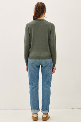 A woman viewed from behind wearing an olive green long-sleeve knit sweater, light blue jeans, and brown sandals against a plain white studio background.