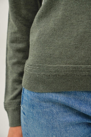 Close-up of a person wearing an olive green ribbed knit sweater with blue jeans, showing the hem and sleeve details.