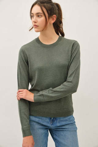 A woman wearing a ribbed long-sleeve crewneck sweater in olive green, paired with blue jeans, photographed against a plain light gray studio background.