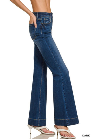 A model wearing dark blue flare jeans with a side pocket and rolled cuffs, paired with white heeled sandals, shown from the waist down on a white background.