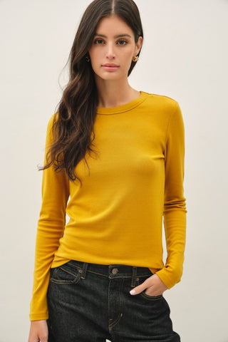 A woman modeling a long-sleeve crewneck knit top in mustard yellow, paired with dark wash jeans, against a plain white background.