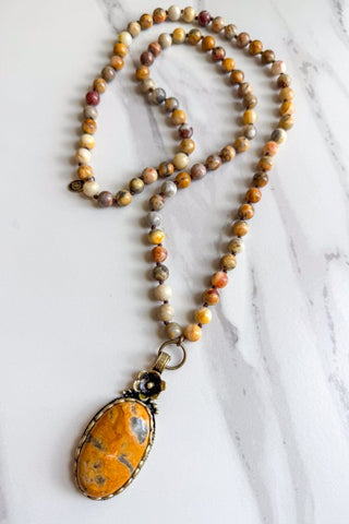 A beaded necklace with a large oval pendant, featuring marbled brown, orange, and cream beads with a prominent orange stone pendant in a silver-toned bezel setting, displayed on a white marble surface.
