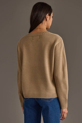A woman shown from the back wearing a beige knit sweater and blue jeans against a plain gray studio background.