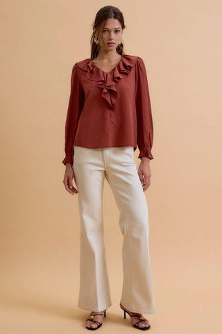 A woman modeling a rust-brown long-sleeve blouse with a ruffled V-neck and cream wide-leg pants against a solid beige studio background.