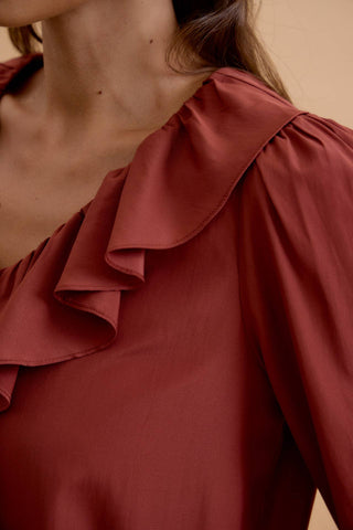 Close-up of a woman wearing a rust-brown satin blouse with a ruffled collar and puffed sleeves against a warm beige background.