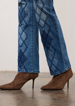 Close-up of a person's legs wearing blue denim jeans with dark blue argyle diamond patterns down the outer sides, paired with brown pointed-toe heeled ankle boots.