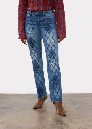 A model wearing blue denim jeans with a dark blue argyle pattern down the front, styled with a red knit sweater and brown high-heeled boots against a neutral studio background.