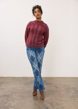 A model stands in a studio wearing a red and blue patterned knit sweater and blue patterned pants with brown ankle boots.